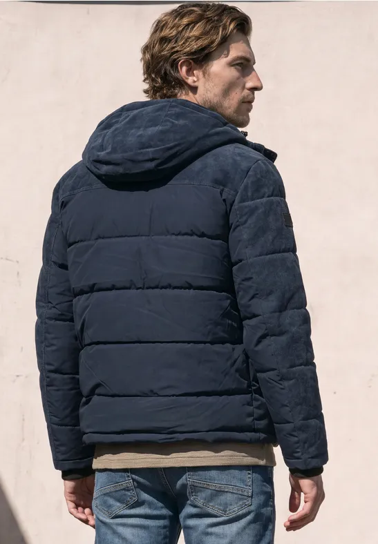 Ein Mann steht in einer blauen Daunenjacke mit Kapuze, Rückenansicht, im Freien vor heller Wand.