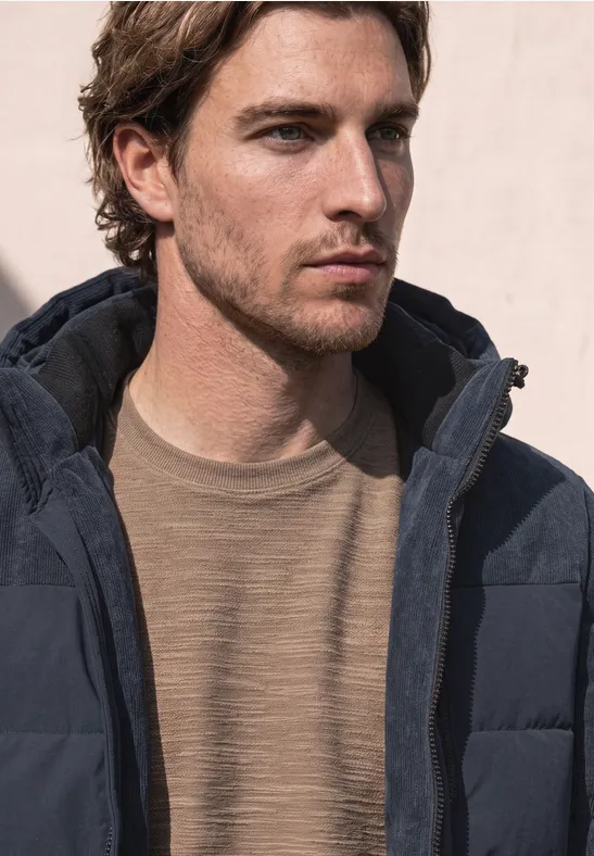 Ein Mann in einer dunklen Steppjacke und beigem Pullover blickt nach rechts vor hellem Hintergrund.