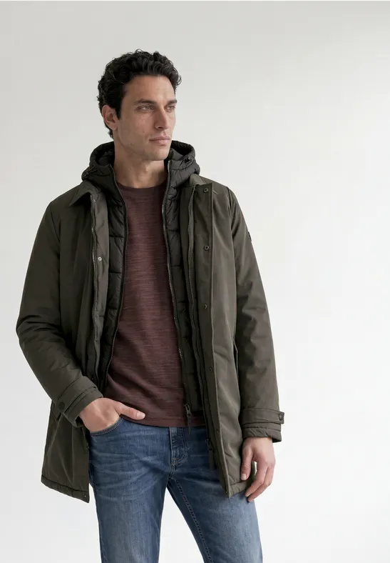 Ein Mann trägt einen dunkelgrünen Parka, Jeans und Pullover und steht vor hellem Hintergrund.