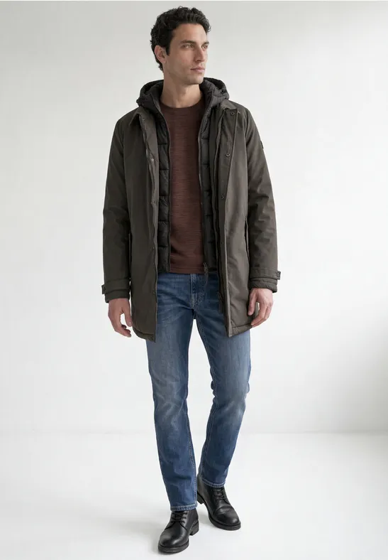 Ein Mann in dunkelgrünem Mantel, rotem Hoodie, blauen Jeans und schwarzen Schuhen vor weißem Hintergrund.