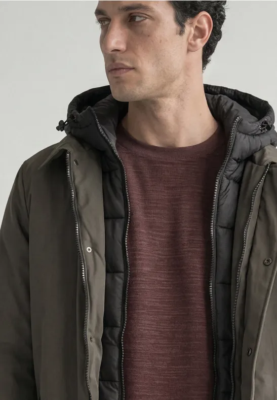 Ein Mann in Profilansicht trägt eine grüne Daunenjacke mit Kapuze und einen bordeauxfarbenen Pullover.