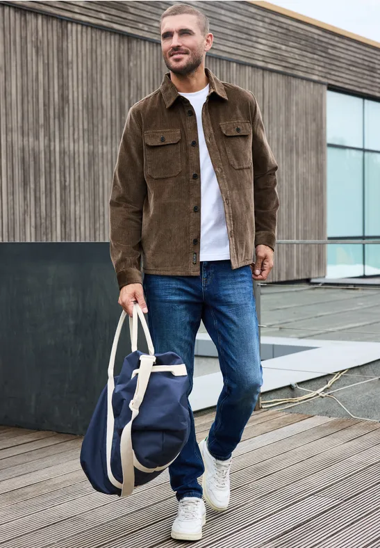 Ein Mann in brauner Jacke und Jeans steht auf einem Holzsteg und hält eine blaue Reisetasche.