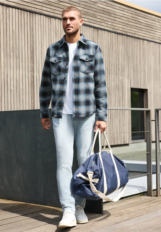 Ein Mann in kariertem Hemd und Jeans trägt eine Reisetasche auf einer Terrasse vor einem Holzhaus.