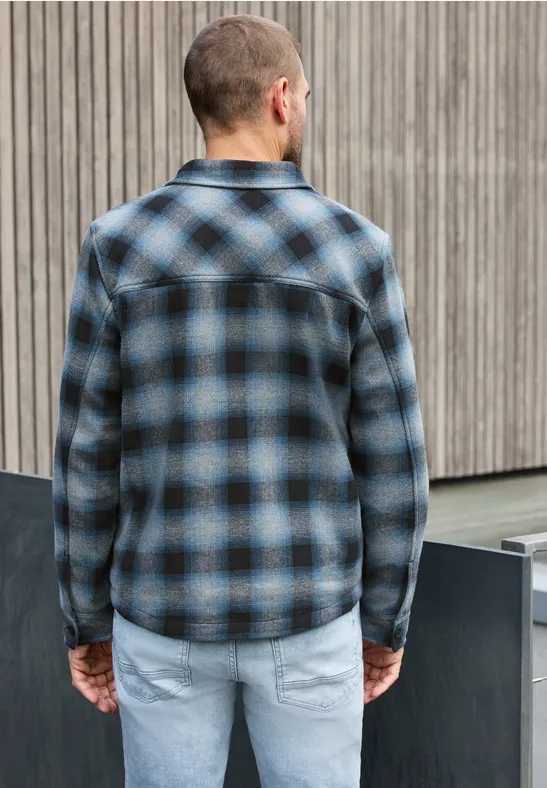 Ein Mann steht von hinten in blau-schwarz kariertem Hemd und hellen Jeans vor einer Holzfassade.