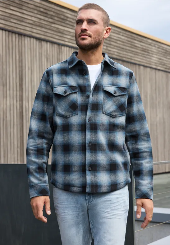 Ein Mann trägt ein kariertes Hemd in Blau- und Grautönen, weißes T-Shirt und helle Jeans vor Holzwand.