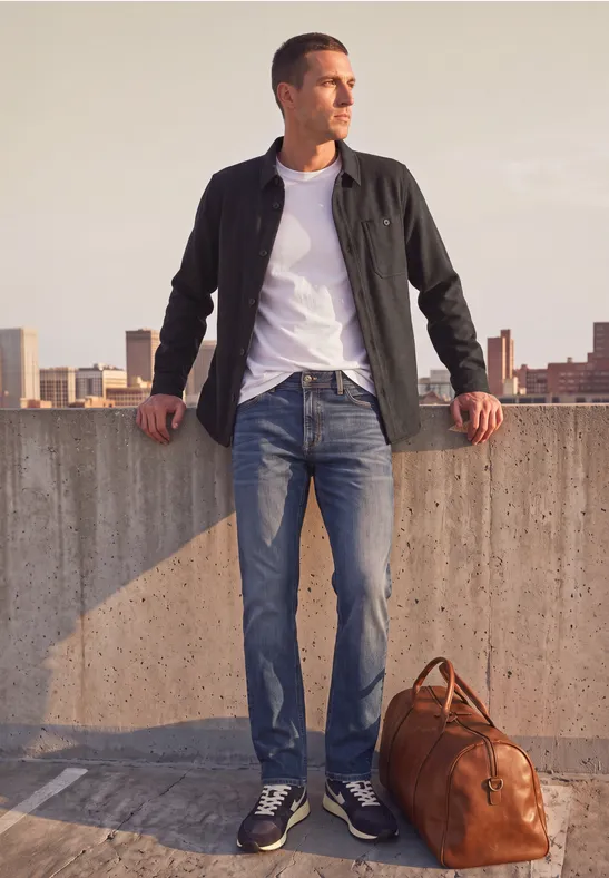 Ein Mann lehnt an einer Betonmauer auf einem Dach, trägt Jeans, Jacke, Sneaker, mit einer Lederreisetasche.