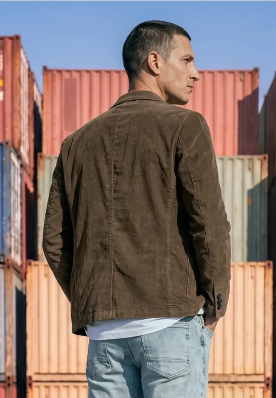 Ein Mann in brauner Jacke und Jeans steht seitlich vor bunten Containern.