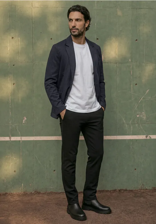 Ein Mann in legerem Outfit (Blazer, weißes T-Shirt, schwarze Hose, Boots) steht vor einer grünen Wand.