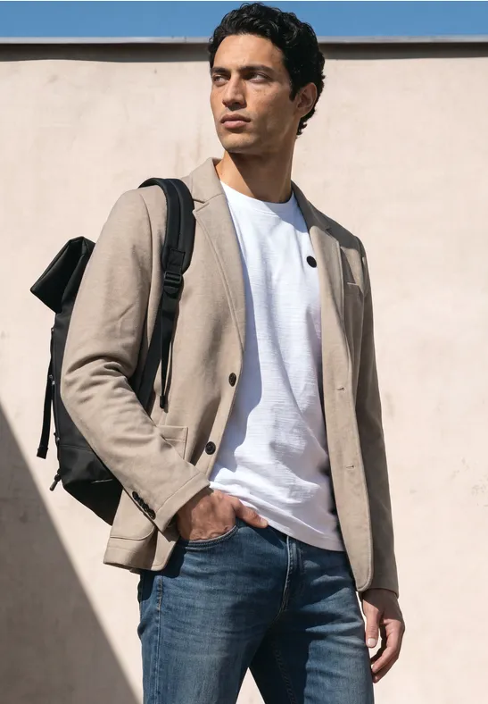 Ein Mann steht vor einer Wand, trägt eine beige Jacke, weißes T-Shirt, blaue Jeans und einen Rucksack.