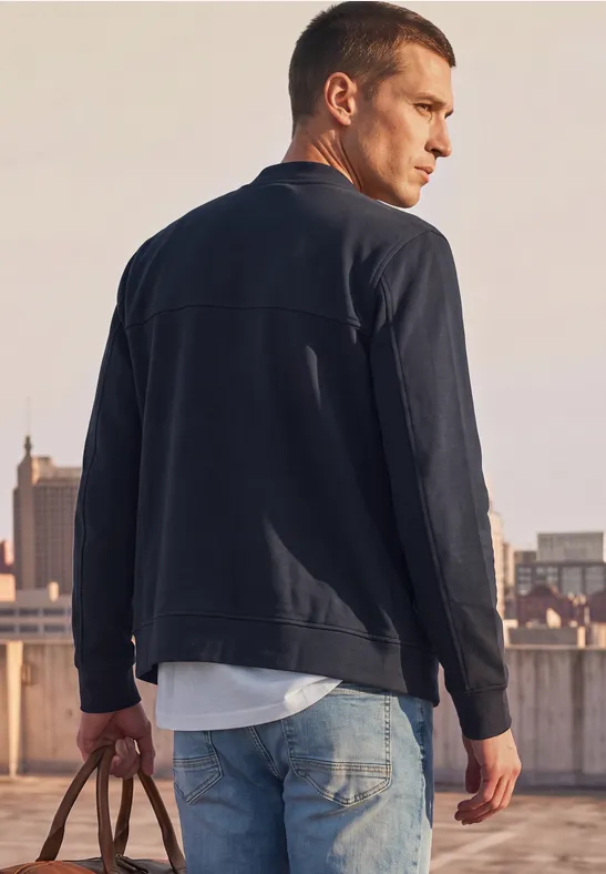 Ein Mann in schwarzer Jacke und Jeans steht auf einem Dach und hält eine Tasche.