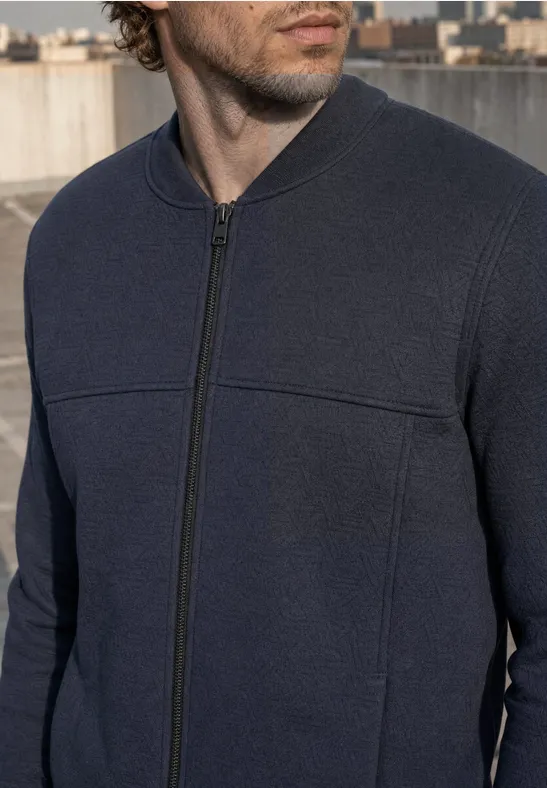 Un homme portant un pull zippé bleu foncé sur un toit avec une ville en arrière-plan.