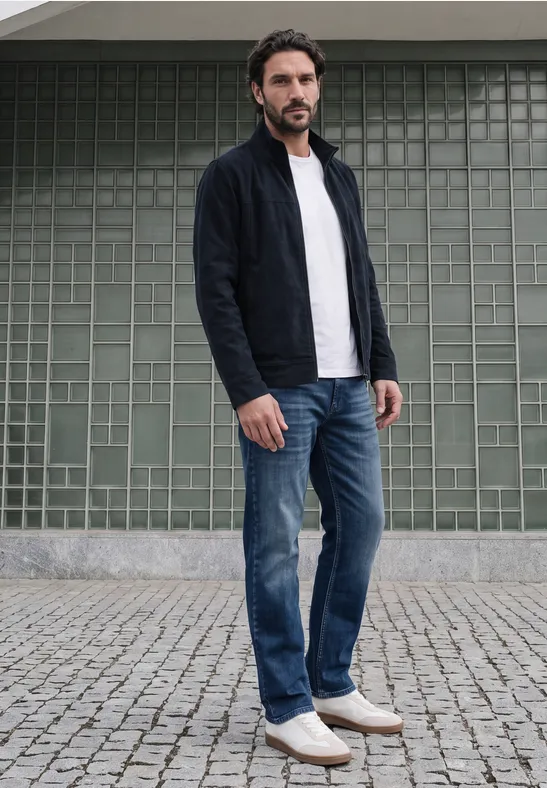 Ein Mann in Jeans, weißem T-Shirt und dunklem Cardigan steht vor einer grünen Fliesenwand auf Kopfsteinpflaster.