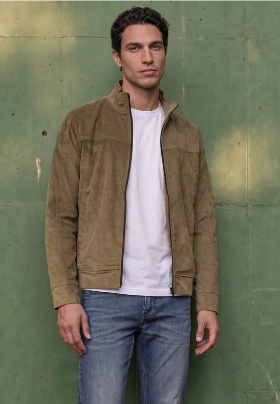 Ein junger Mann in brauner Cord-Jacke, weißem T-Shirt und Jeans steht vor einer grünen Wand.