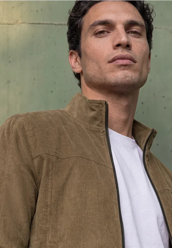 Junger Mann in brauner Cordjacke mit hochgeschlossenem Kragen, weißem T-Shirt, vor grünlicher Wand.