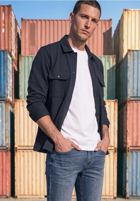 Un homme en jean, T-shirt blanc et veste noire pose décontracté devant des conteneurs colorés.