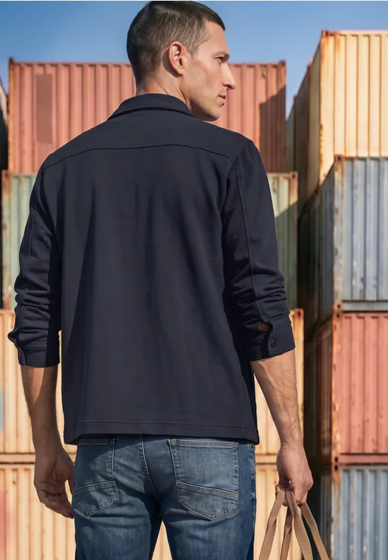 Un homme vu de dos en veste sombre et jean se tient devant des containers colorés, tenant un sac.