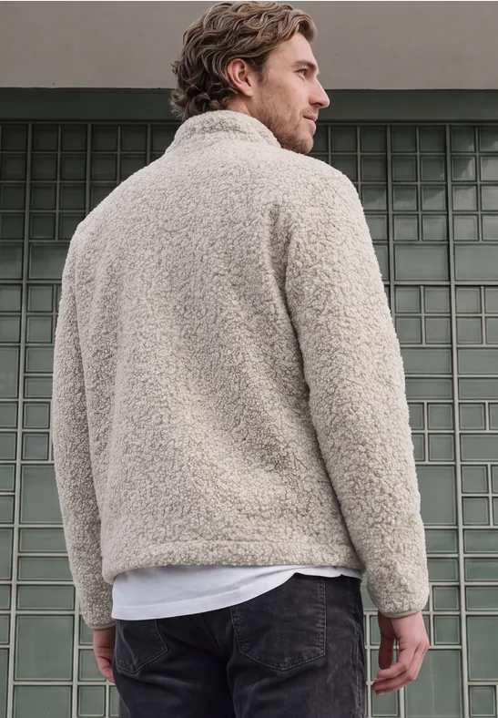 Ein Mann trägt eine beige Kuscheljacke und steht mit dem Rücken zur Kamera vor einer Mauer.