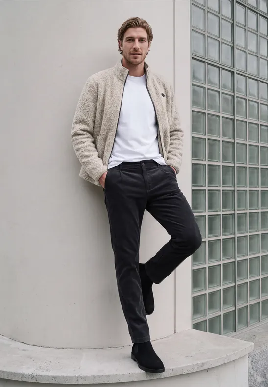 Mann lehnt lässig an weißer Wand, trägt Jacke, weißes Shirt und dunkle Hose, Blick zur Kamera.