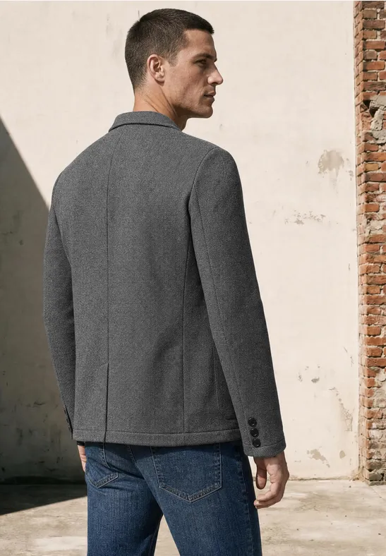 Ein Mann steht von hinten, trägt grauen Blazer und blaue Jeans vor einer hellen Wand.