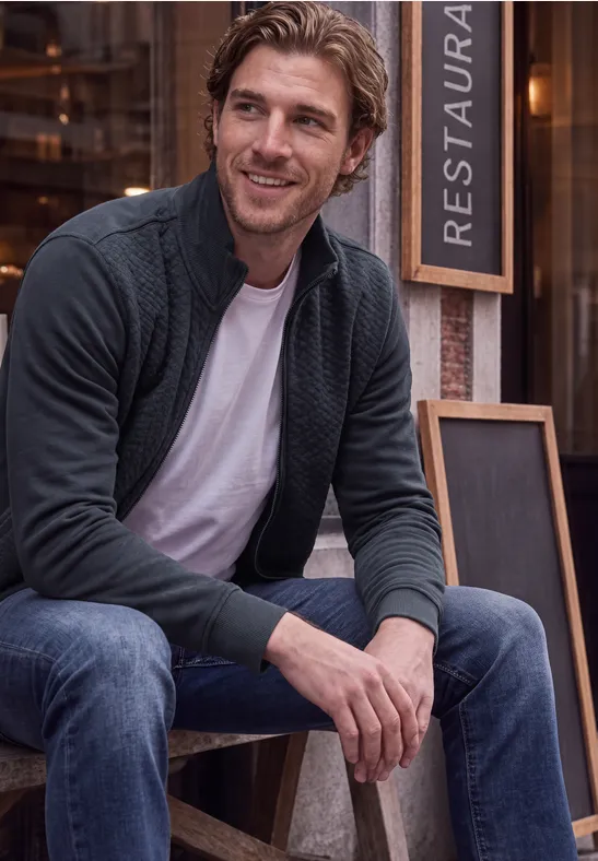 Ein lächelnder Mann sitzt vor einem Restaurant, trägt Jeans, T-Shirt und Strickjacke.