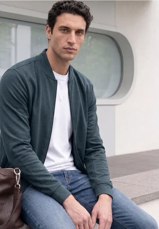 Ein Mann sitzt auf einer Bank, trägt grüne Jacke, weißes T-Shirt, blaue Jeans und hat eine braune Ledertasche.