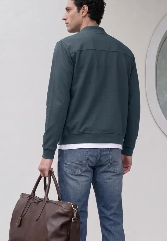 Mann von hinten in Jeans, grauer Jacke, trägt eine braune Ledertasche vor weißer Wand mit rundem Fenster.