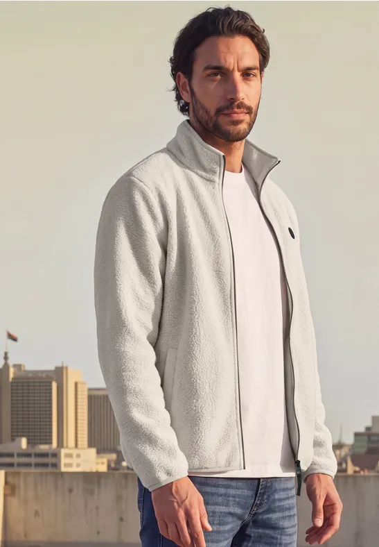 Ein Mann trägt eine graue Fleecejacke über weißem T-Shirt, stehend auf einem städtischen Dach.