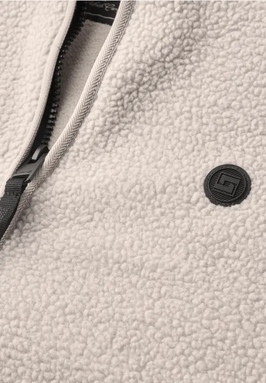 Nahaufnahme einer cremefarbenen Fleecejacke mit Reißverschluss und rundem Markenlogo