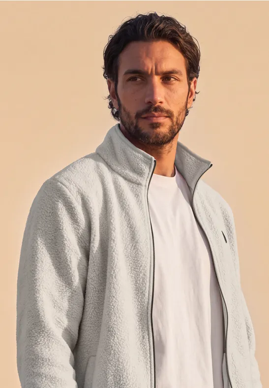 Ein Mann mit dunklen Haaren und Bart trägt eine graue Fleecejacke und weißes T-Shirt vor hellem Hintergrund.