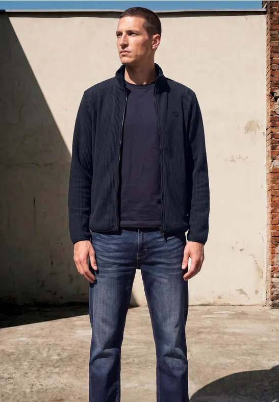 Ein Mann in Jeans, T-Shirt und Jacke steht vor einer hellen Wand im Freien.
