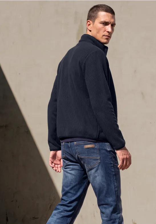Ein Mann in dunkler Jacke und Jeans blickt über die Schulter vor einer hellen Wand.