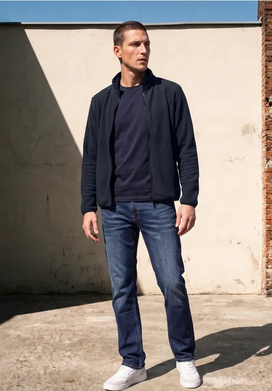 Ein Mann steht im Sonnenlicht vor einer Wand, trägt blaue Jeans, weiße Sneaker, navy T-Shirt und Jacke.