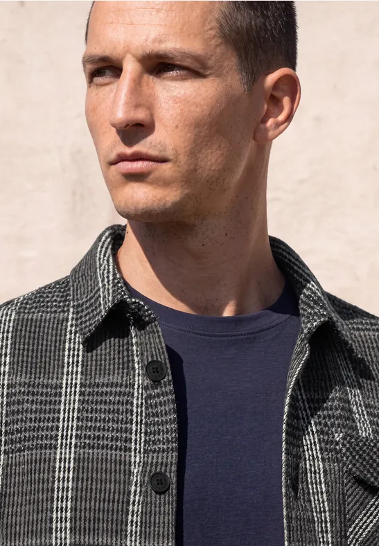 Ein ernster Mann in kariertem Hemd und dunkelblauem Shirt blickt seitlich.