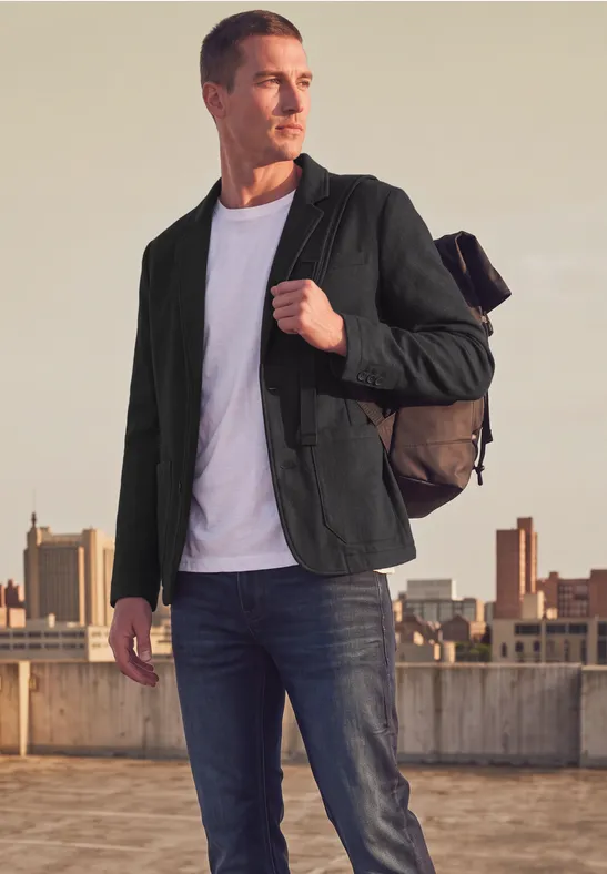 Mann mit Rucksack, grüner Jacke, weißem Shirt und Jeans steht auf einem Dach vor einer Stadtkulisse.