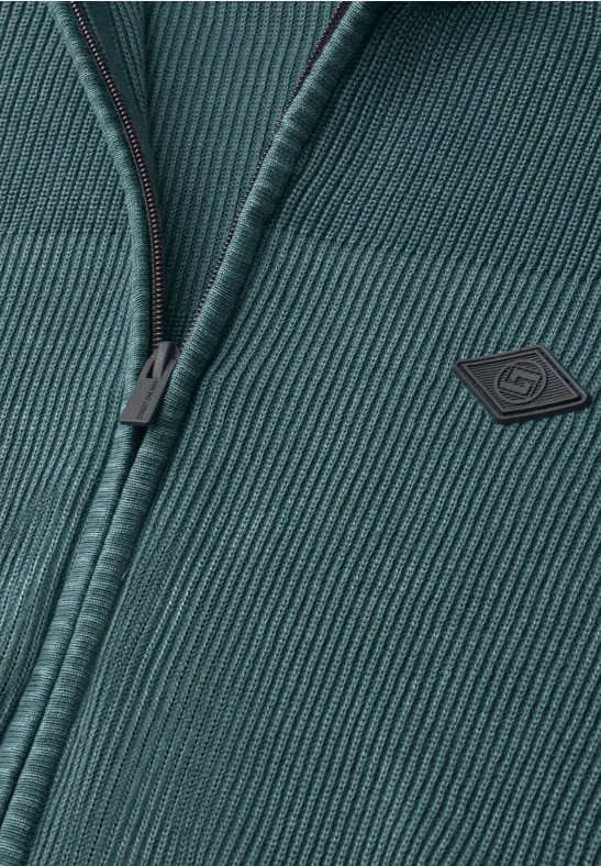 Close-up van een donkergroene, geribbelde gebreide jas met rits en klein logo op de borst.