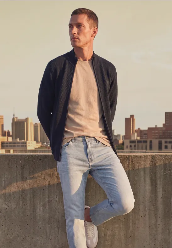 Ein Mann in Jeans, T-Shirt und Jacke steht lässig auf einem Dach vor einer urbanen Skyline.