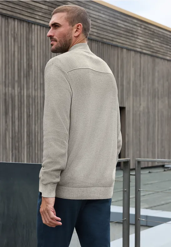 Ein Mann steht auf einem Dach vor einer Holzfassade, trägt eine graue Strickjacke und schaut seitlich.