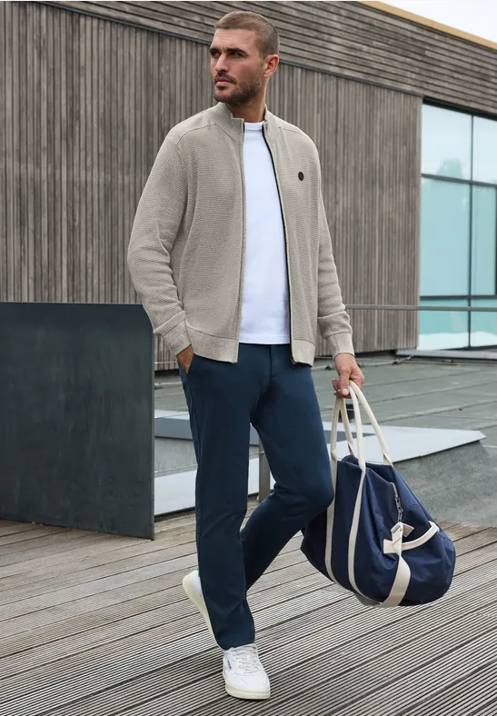 Ein Mann in grauer Jacke und dunkler Hose spaziert mit Tasche auf einem Holzsteg.