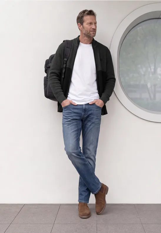 Un homme décontracté appuyé contre un mur près d'une fenêtre ronde, vêtu de jean, tee-shirt blanc, veste et sac à dos.