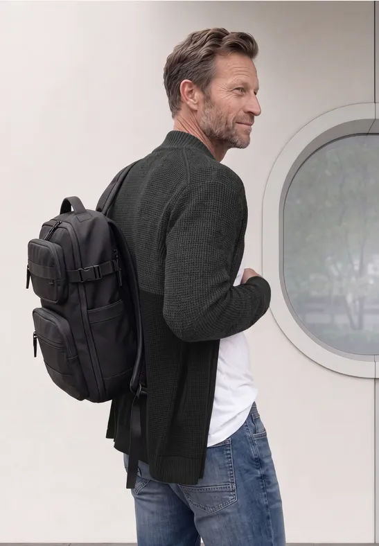 Un homme en jean et blazer porte un sac à dos noir et regarde sur le côté devant un mur blanc avec une fenêtre ronde.