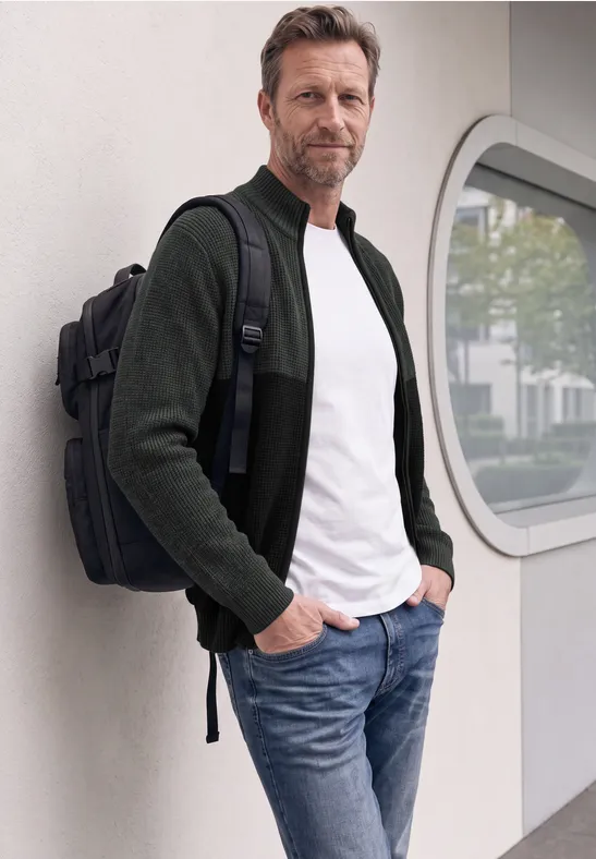 Un homme en t-shirt blanc, veste foncée et jean, avec un sac à dos, appuyé contre un mur blanc.