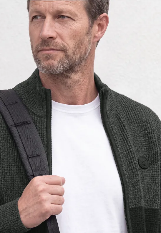 Un homme d'âge moyen à la barbe grise porte un gilet vert, un T-shirt blanc et un sac bandoulière.