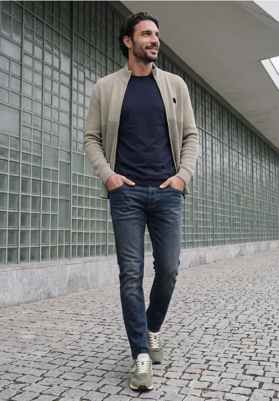 Mężczyzna z brodą w beżowej kurtce, granatowym t-shircie i dżinsach idzie po brukowanym chodniku.