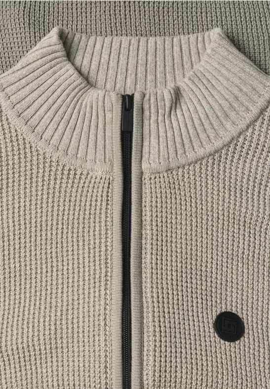 Beżowy sweter z grubego splotu, stójką, centralnym zamkiem i małą czarną okrągłą naszywką z przodu.