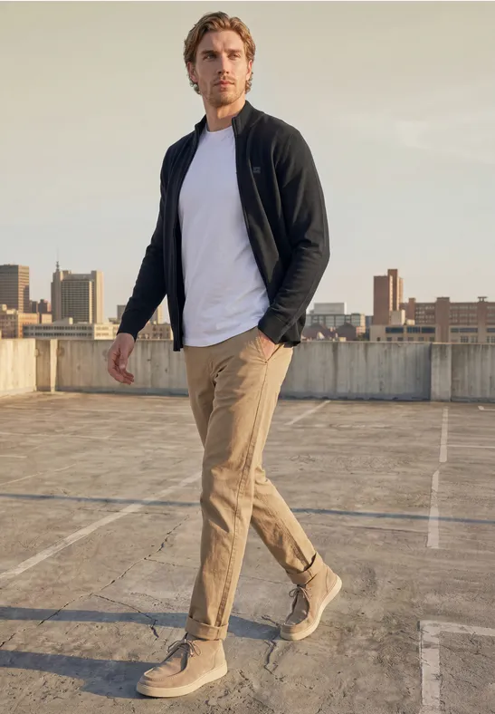 Ein Mann in weißem T-Shirt, schwarzer Jacke und khakifarbenen Hosen geht auf einem Parkdeck mit Stadthintergrund.