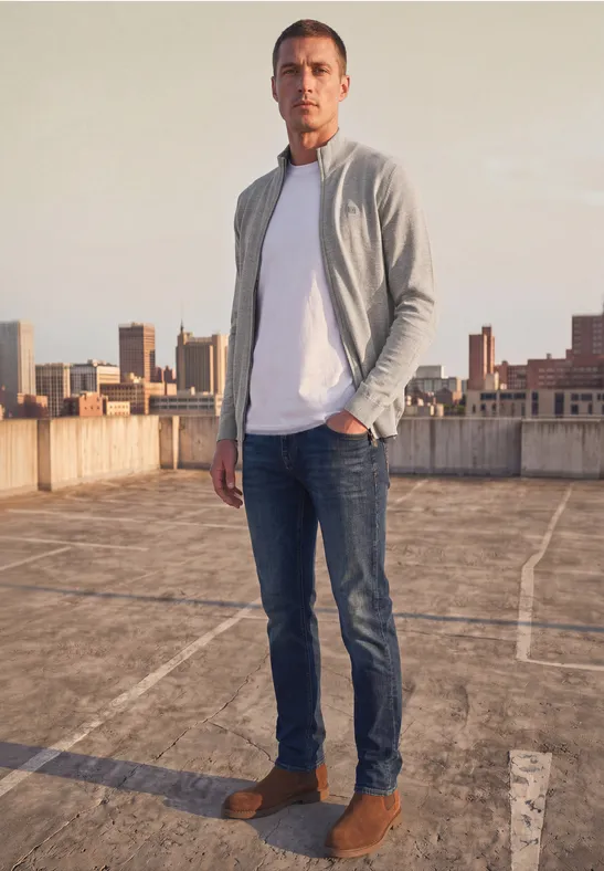 Ein Mann steht auf einem Dachparkplatz vor einer Stadtkulisse, trägt Jeans, weiße T-Shirt, graue Jacke und braune Schuhe.