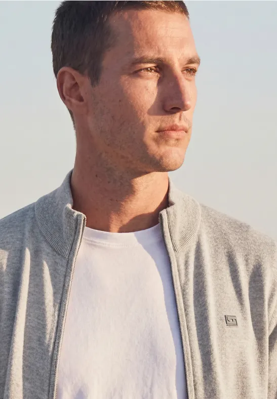 Ein junger Mann steht im natürlichen Licht und trägt ein weißes T-Shirt und eine graue Jacke.
