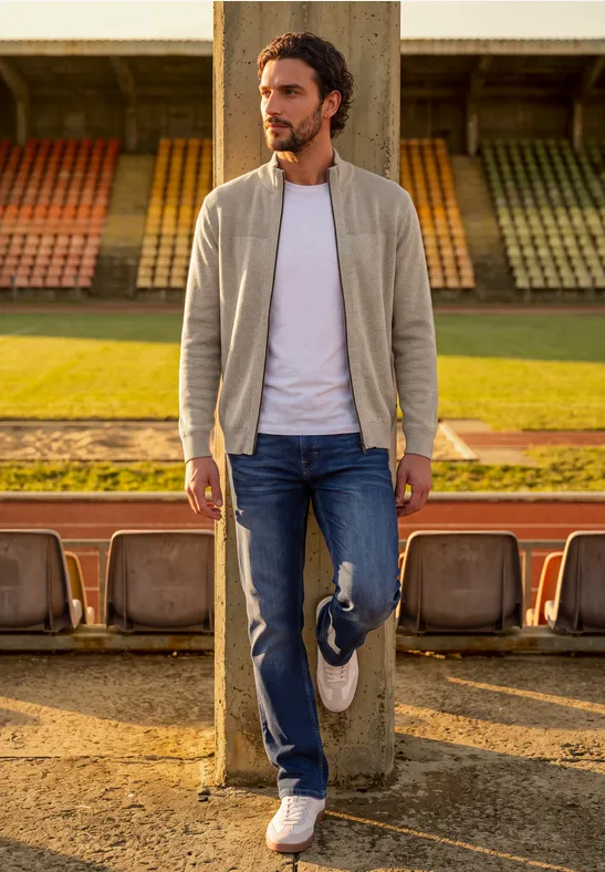 Ein Mann lehnt lässig an einer Säule auf der Tribüne, trägt Bomberjacke, Jeans und hält Turnschuhe.