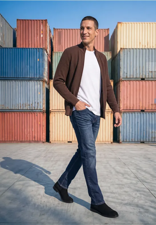 Ein Mann in Jeans, weißem T-Shirt und brauner Jacke geht vor bunten Containern spazieren.