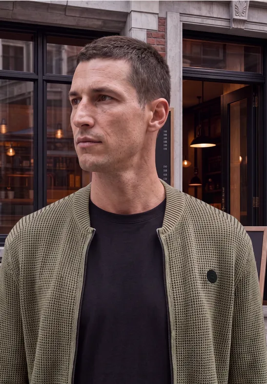Ein Mann mit kurzem Haar trägt eine olivgrüne Strickjacke und schaut zur Seite.